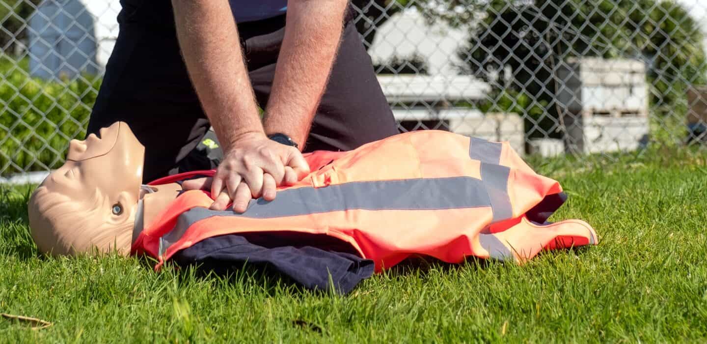 Person performing CPR chest compressions on a training mannequin outdoors on grass.