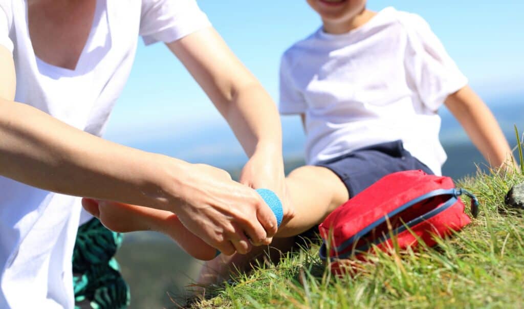 Person providing first aid by wrapping a bandage on a child's foot outdoors during a hike.