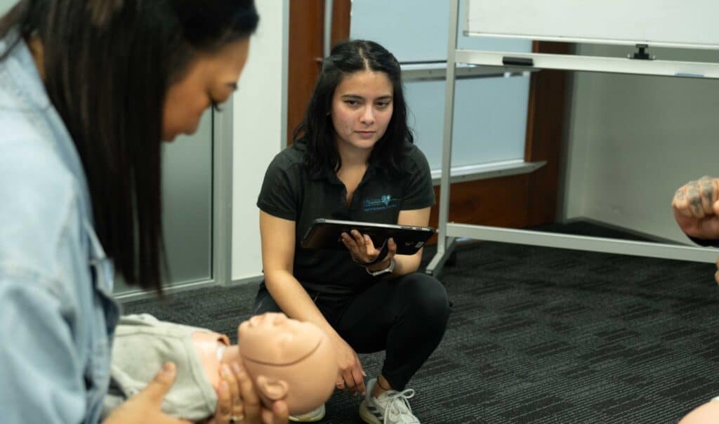 Instructor observes first aid training session with infant CPR manikin in classroom setting.